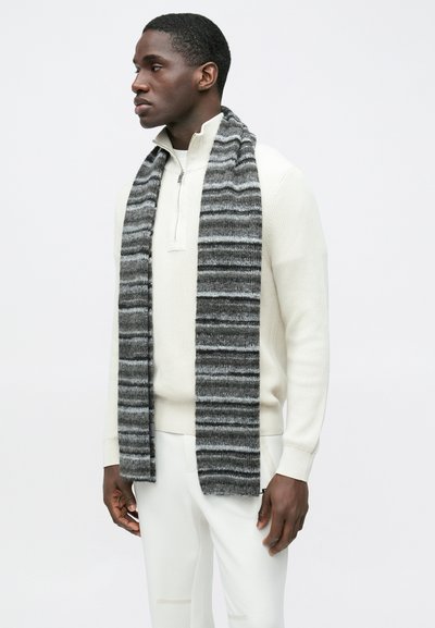 Ριγέ κασκόλ σε γκρι και μαύρο πλεκτό, με απαλό υφασμάτινο, κρέμεται δίπλα σε κρεμ πουλόβερ. Στενή εφαρμογή με φερμουάρ.