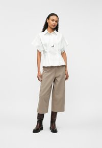 Λευκή κοντομάνικη μπλούζα με ζώνη peplum και μαύρες λεπτομέρειες, συνδυασμένη με καφέ παντελόνια φαρδιάς γραμμής και καφέ μπότες μέχρι το γόνατο.