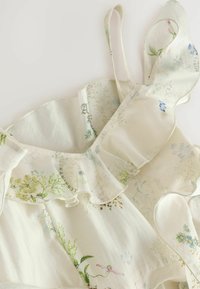 Close-up af hvid blomsterkjole med flæsekant og tynde skulderstropper på en lys baggrund.