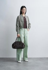Femme portant un cardigan en maille gris, un haut marron, un pantalon large vert clair, des chaussures blanches pointues, tenant un grand sac à main marron foncé.