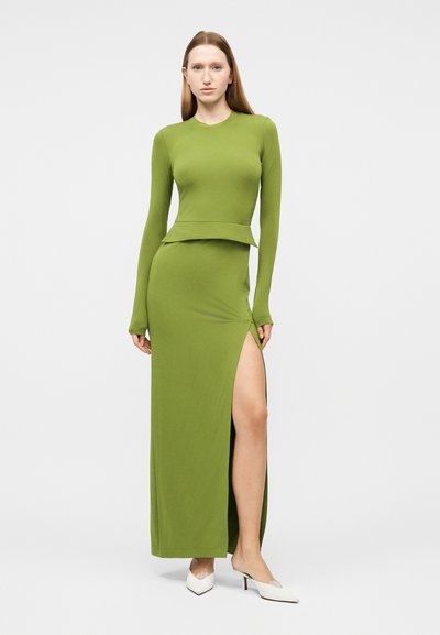 Γυναίκα που φοράει μακρυμάνικο φόρεμα σε ελιά πράσινο χρώμα με ψηλή σχισμή στην αριστερή πλευρά, σε συνδυασμό με λευκές μυτερές γόβες.