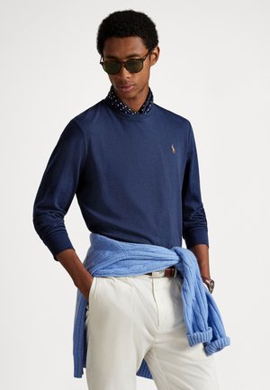 Temno modra polo majica z dolgimi rokavi in rebrastim ovratnikom, kombinirana s svetlo modrim puloverjem, pripetim okoli pasu. Na vratu je viden okrogel vzorec s pikami.