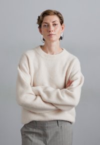 Svetlá krémová pletená mikina s kulatým výstrhem, v kombinácii so šedými pruhovanými nohavicami. Model má prekrížené ruky a veľké strieborné náušnice.