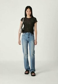 Νεαρή γυναίκα που στέκεται, φορώντας μαύρη δαντελένια μπλούζα, ανοιχτό γαλάζιο τζιν, μαύρα σανδάλια και κρατώντας μία ασημένια τσάντα ώμου, σε λευκό φόντο.