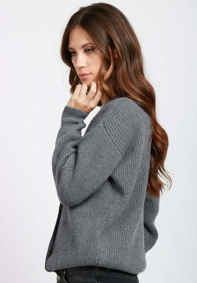 Donna con lunghi capelli castani che indossa un maglione grigio a coste, toccandosi il mento, con uno sguardo pensieroso su uno sfondo chiaro semplice.