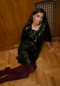 Fekete flitteres ruha, laza szabással és rövid ujjakkal. A modell bordó harisnyát visel, és egy fa falnak támaszkodik, kiemelve a ruha textúráját és fényét.