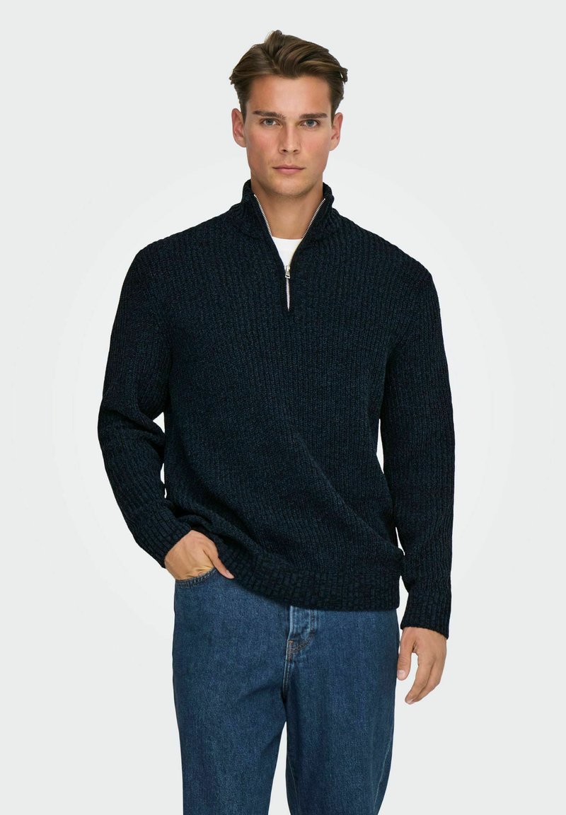 Navy-blauer Strickpullover mit hohem Kragen und Reißverschluss, mit einem strukturierten Muster. Kombiniert mit hellblauen Jeans.