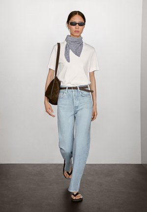 White t-shirt, light blue jeans, patterned scarf, brown suede bag, black sandals. Plain background, model walking.
