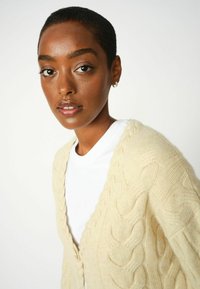 Cremefarbener Kabelstrick-Cardigan mit einem strukturierten Muster, offener Vorderseite und weichem Stoff, kombiniert mit einem weißen Rundhalsausschnitt-T-Shirt.