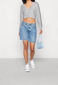 Szara krótka kardigan z teksturowanym wzorem, zestawiona z jasnoniebieskimi szortami dżinsowymi i białymi trampkami. Trzyma małą niebieską torebkę.