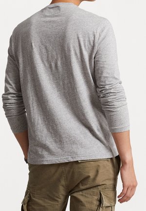 Personne portant une chemise à manches longues gris clair et un pantalon cargo vert olive, vue de dos sur un fond uni.