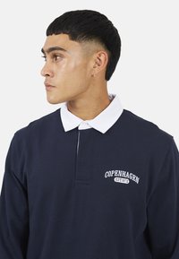 Polo a maniche lunghe blu navy con colletto bianco, con ricamo "COPENHAGEN EST. 1972" sul petto, realizzato in una morbida miscela di cotone.