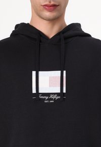 Felpa nera con un motivo frontale a strisce blu chiaro, bianche e rosse. Presenta un logo centrale in bianco con la scritta "Tommy Hilfiger EST. 1985."