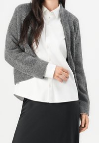 Szary wełniany kardigan o luźnym kroju, z długimi rękawami i ściągaczami, noszony na białej koszuli z guzikami i warstwowo na czarnej spódnicy.