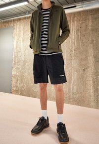 Tmavě zelená fleecová bunda, pruhované šedé tričko, černé síťované šortky a černé boty s gumovou podrážkou. Pod šortkami jsou vidět bílé ponožky.