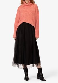 Maglione oversize in corallo lavorato a maglia con collo a imbuto, abbinato a una gonna midi plissettata in tulle nera. Stivali neri alla caviglia con tacco a blocco.