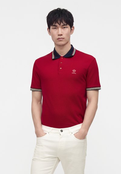 Homme portant un polo rouge Versace avec col noir et pantalon blanc, debout les mains dans les poches sur un fond uni.