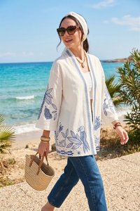 Woman walking on beach path wearing white embroidered jacket, blue jeans, sunglasses, headband, and carrying a woven straw handbag.