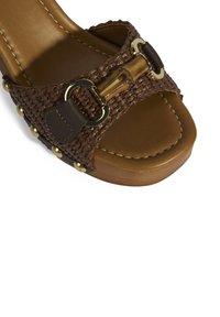 Braune geflochtene Rutsch-Sandale mit einem glänzenden braunen Riemen, goldenen Hardware-Details und einer strukturierten Kunstleder-Sohle. Verfügt über ein Plattform-Design.