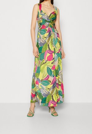 Femme portant une robe sans manches, à encolure en V, longue jusqu’au sol, avec des motifs de feuilles multicolores éclatants et des sandales à talons hauts vertes à lanières.