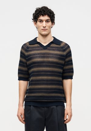 Jeune homme aux cheveux bouclés foncés portant un polo en maille à manches courtes rayé bleu marine et marron et un pantalon foncé sur fond blanc.
