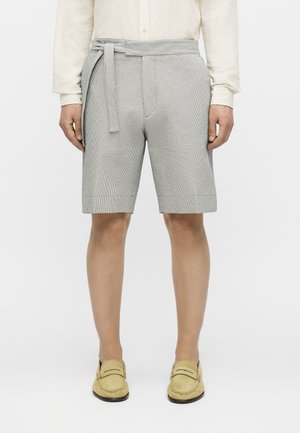 Muž nosi šedé textúrované šortky s vysokým pásom a látkovým opaskom, béžovú košeľu s dlhým rukávom a svetložlté mokasíny, stojí na bielom pozadí.