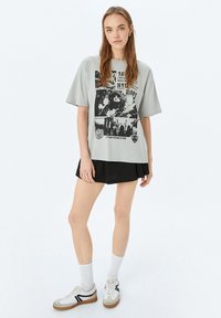 Lichtgrijze oversized t-shirt met zwarte grafische prints, korte mouwen, gecombineerd met een zwarte rok en witte sneakers met grijze en zwarte accenten.