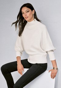 Weißer, strukturierter Rollkragenpullover mit weiten Ärmeln, kombiniert mit schwarzen, schmal geschnittenen Hosen. Modell sitzt auf einem weißen Block und trägt hängende Ohrringe.