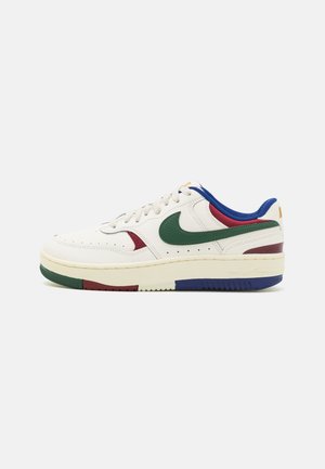 Hvid lavtop sneaker med grøn Nike swoosh, røde og blå accenter, creme sål og snørelukning, vist mod en hvid baggrund.