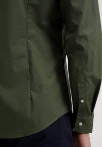 Zbliżenie na osobę noszącą ciemnozieloną, zapinaną na guziki, koszulę z długim rękawem oraz granatowe spodnie, pokazujące detale mankietu i dolnego wykończenia koszuli.