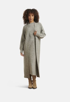 Donna in piedi che indossa un lungo cardigan grigio lavorato a maglia e un abito abbinato con stivali al ginocchio in suede beige, su uno sfondo bianco.