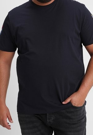 Mężczyzna ubrany w prostą czarną koszulkę z krótkim rękawem i ciemne dżinsy, z jedną ręką w kieszeni, a drugą opuszczoną wzdłuż ciała.