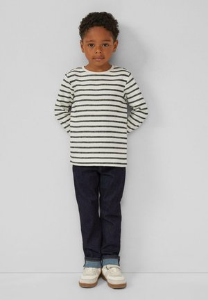 Gestreepte lange mouwen shirt in crème en donkergrijs, gecombineerd met donkerblauwe jeans. Witte sneakers met beige accenten. Kind dat met handen achter de rug staat.