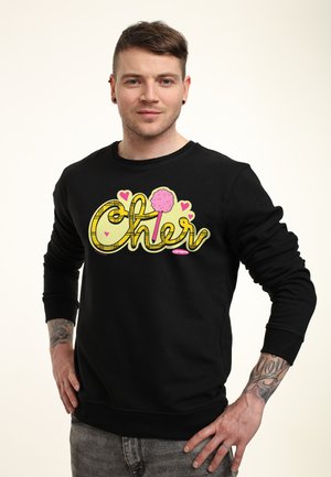 Mand med kort hår og tatoveringer iført en sort sweater med farverigt "Clueless"-logo og hjerter, står med hænderne på hoften.