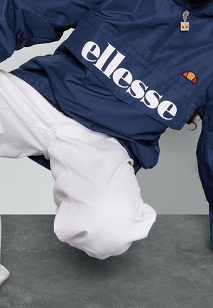 Person iført navy blå ellesse jakke og hvide bukser, placeret i en krummende position mod en grå baggrund.