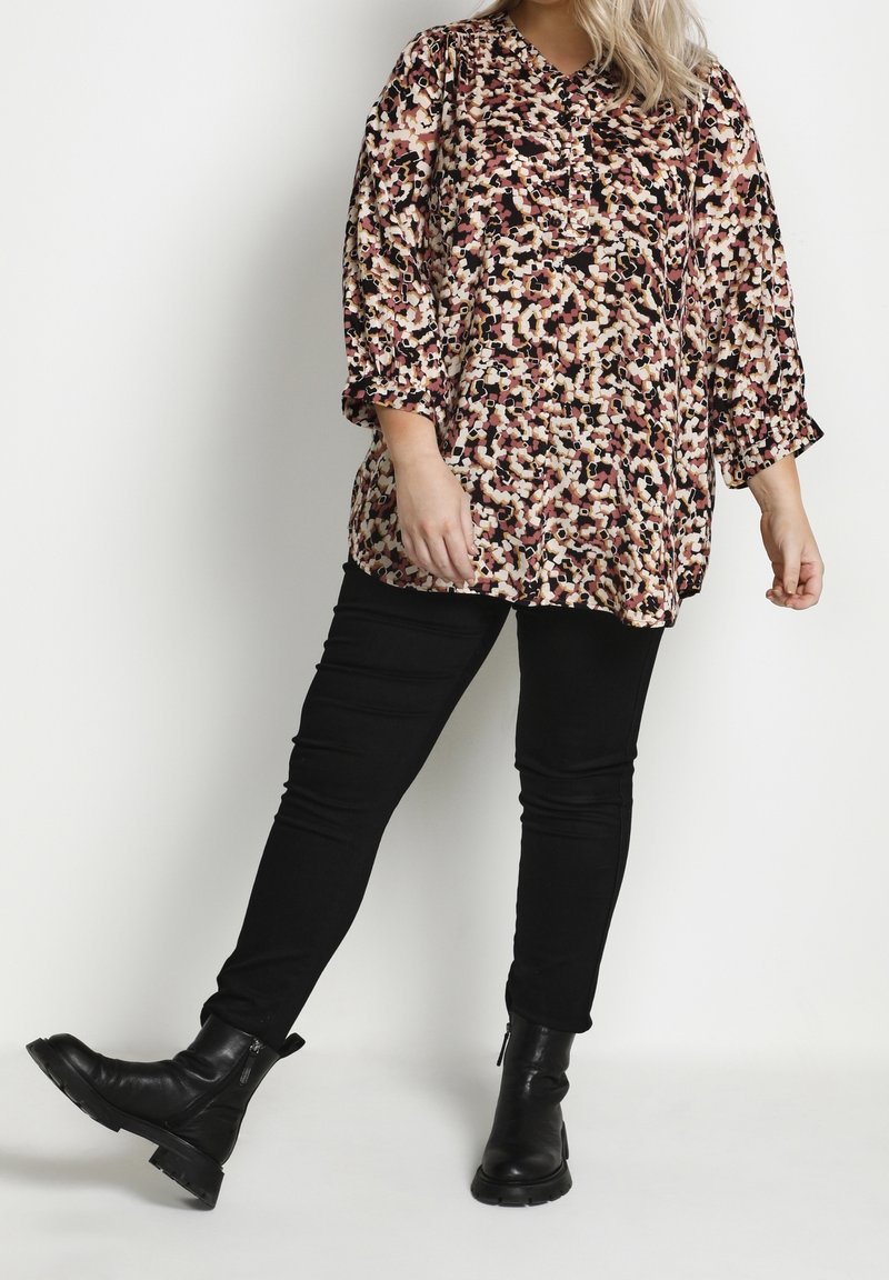 Femme portant un pantalon noir, des bottes noires et un chemisier ample à motif floral avec des manches trois quarts, debout devant un fond clair uni.