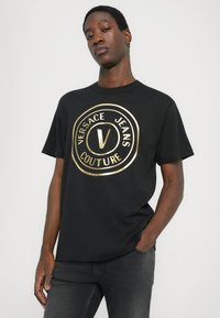 Tricou din bumbac negru cu un logo rotund auriu care include "VERSACE JEANS COUTURE" și un design central cu litera "V". Mâneci scurte, croială lejeră.