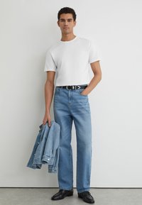 Mand iført hvid T-shirt, blå jeans, sort bælte og sorte sko, der holder en blå denimjakke i venstre hånd.