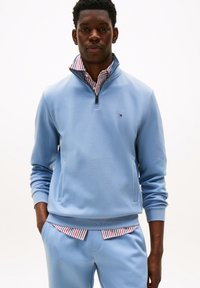 Sweatshirt zippé bleu clair avec des poignets côtelés, comportant une poche avant et un col à rayures blanches et rouges dépassant dessous.