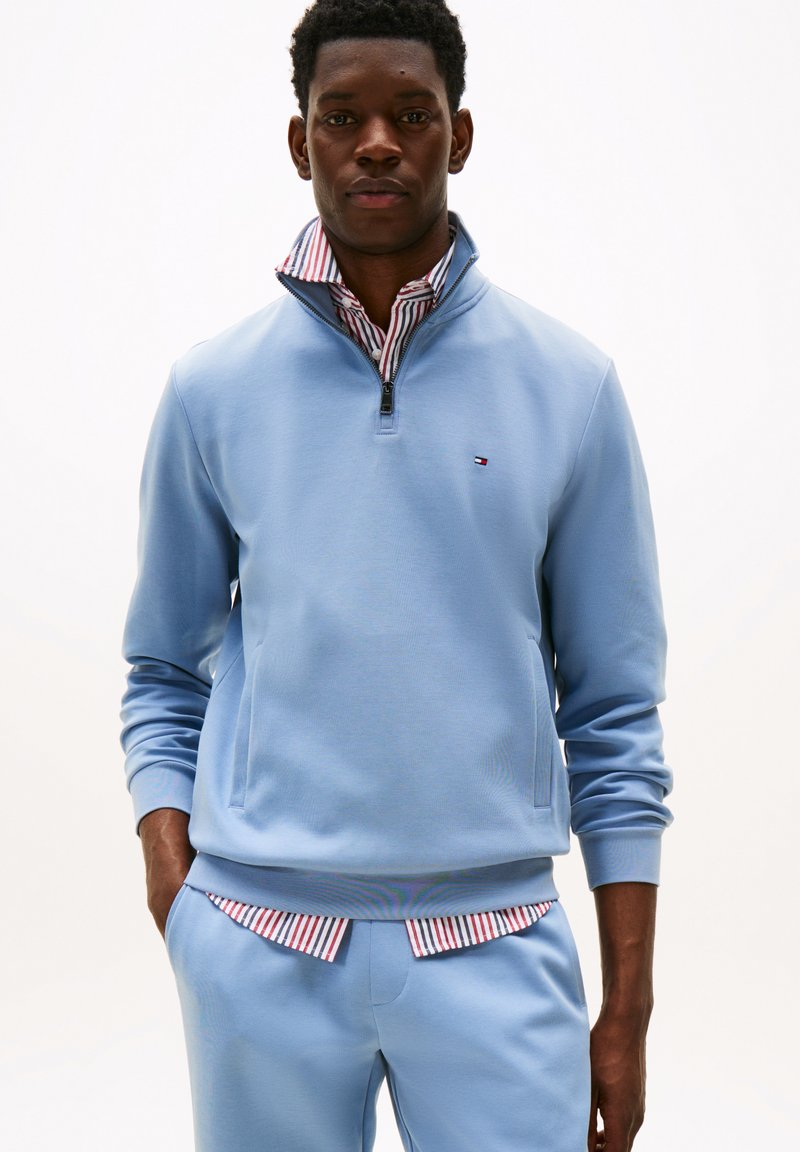 Sweatshirt zippé bleu clair avec des poignets côtelés, comportant une poche avant et un col à rayures blanches et rouges dépassant dessous.