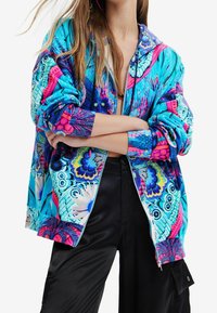 Kolorowa kurtka bomberka z żywymi wzorami w odcieniach niebieskiego, różowego i fioletowego. Wykonana z gładkiego materiału, zapinana na zamek błyskawiczny, z prążkowanymi mankietami.