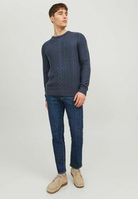 Mørkeblåt cable knit sweater med rund halsudskæring, struktureret mønster, ribstrikkede ærmer og søm. Kombineret med blå jeans og lyse sko.