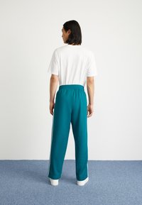 Teal joggebukser med hvite side-striper og elastisk linning, kombinert med en enkel hvit t-skjorte, sett fra baksiden.