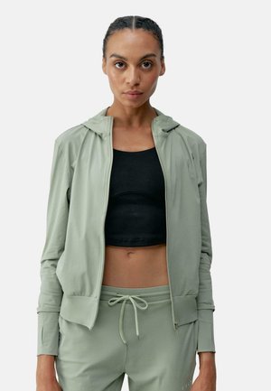 Ljusgrön zip-up hoodie med huva, figurnära design och ribbade muddarna, i kombination med en svart crop top och matchande byxor. Slät textur.