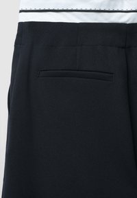 Pantaloni di un blu scuro realizzati in tessuto testurizzato, con una tasca posteriore singola. La vita è strutturata e liscia, con cuciture visibili.