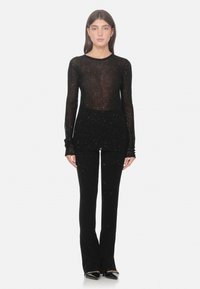 Vrouw in een doorschijnende zwarte blouse met lange mouwen en bijpassende zwarte broek met subtiele glitters, staand met het gezicht naar voren op een witte achtergrond.