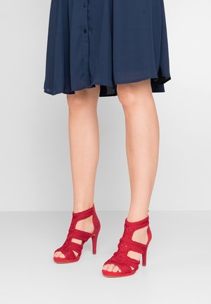 Jambes vêtues d'une robe bleu marine et de sandales à talons hauts rouges avec des brides tressées et des orteils ouverts, se tenant sur un sol gris clair.