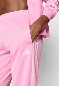 Ροζ αθλητικό παντελόνι από λείο ύφασμα με τρεις λευκές ρίγες και λογότυπο Adidas. Διαθέτει ελαστική μέση και πλαϊνές τσέπες.