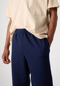 Tricou bej din amestec de bumbac, asortat cu pantaloni jogger bleumarin, având un tiv elastic și buzunare laterale, evidențiind un stil casual și o textură moale.