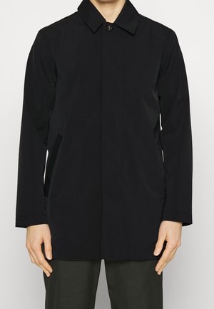 Mann trägt einen schwarzen, langärmeligen, geknöpften Mantel mit Seitentaschen und steht vor einem schlichten Hintergrund.